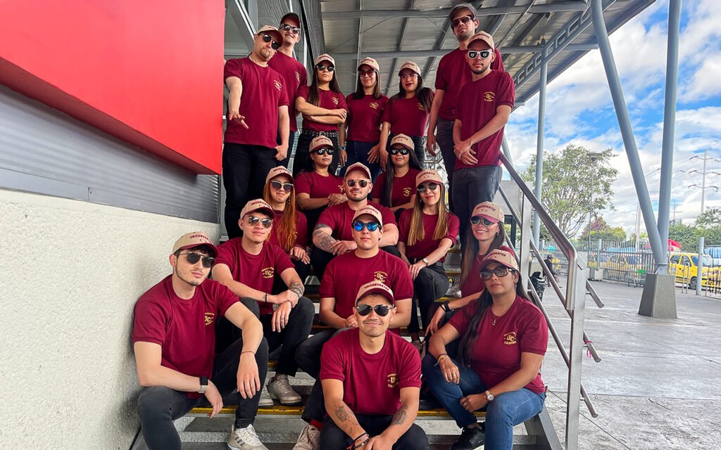 Equipo de UNICERVANTES con camisetas burdeos y gorras beige, posando en las escaleras del Portal Norte durante el evento de alianza con TransMilenio.