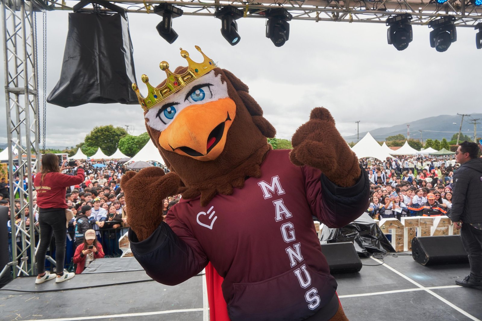 La mascota universitaria “Magnus”, con una corona dorada, posa animadamente desde el escenario principal del Brunch Fest 2025, mientras una multitud de estudiantes disfruta del evento en el fondo.